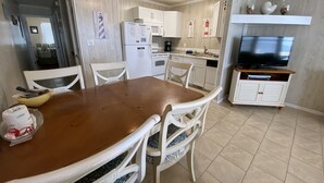 Een compacte keuken met witte kasten, een eettafel met stoelen, een plafondventilator en een televisie op een standaard.