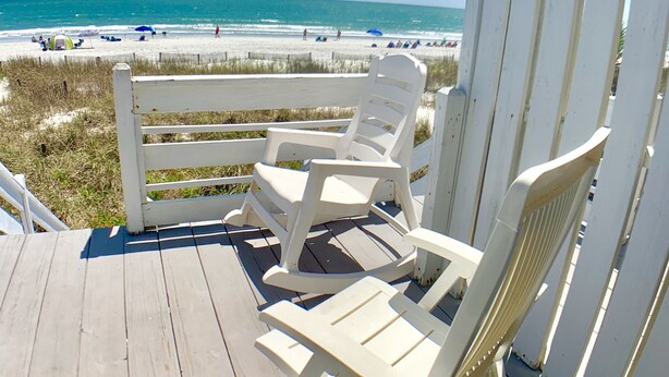 Een houten terras met witte Adirondack-stoelen met uitzicht op een strand.