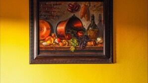 Pintura emoldurada de natureza morta com frutas e legumes numa parede amarela.