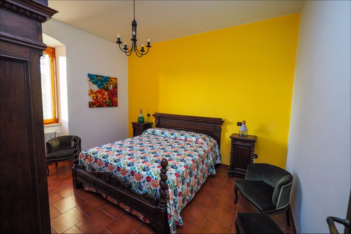 A bedroom with a floral bedspread, a wooden headboard, a nightstand, a chair, and a painting on the wall.