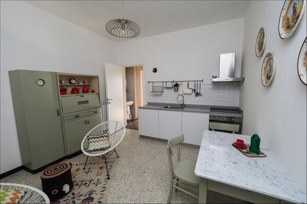 A compact kitchen with a dining area, a green cabinet with tea sets, a white chair, and a marble countertop.