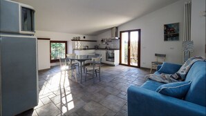 Uma sala ampla com um sofá azul, uma mesa de jantar com cadeiras, uma cozinha com fogão e pia, e uma varanda com vista.