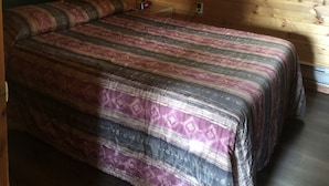 Ein Zimmer in einer Holzhütte mit einem Bett, einem Nachttisch, einer Lampe und einem Fenster mit Vorhängen.