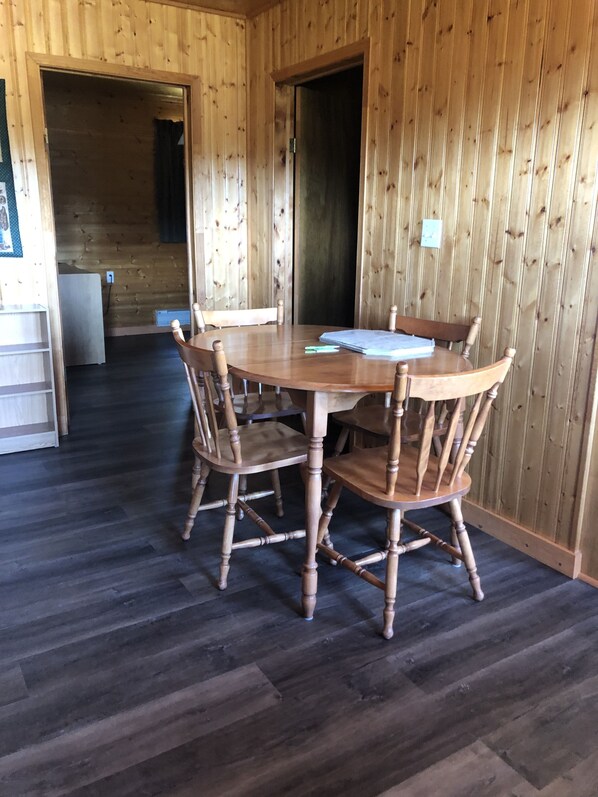 Interior de madera con una mesa redonda y cuatro sillas, un refrigerador y una puerta que da a otra habitación.