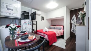 A compact hotel room with a bed, a dining area with a table set for two, a microwave, a small refrigerator, and a TV. - North Cottage | Shibuya Studio | Close to Meiji Shrine (Shibuya)
