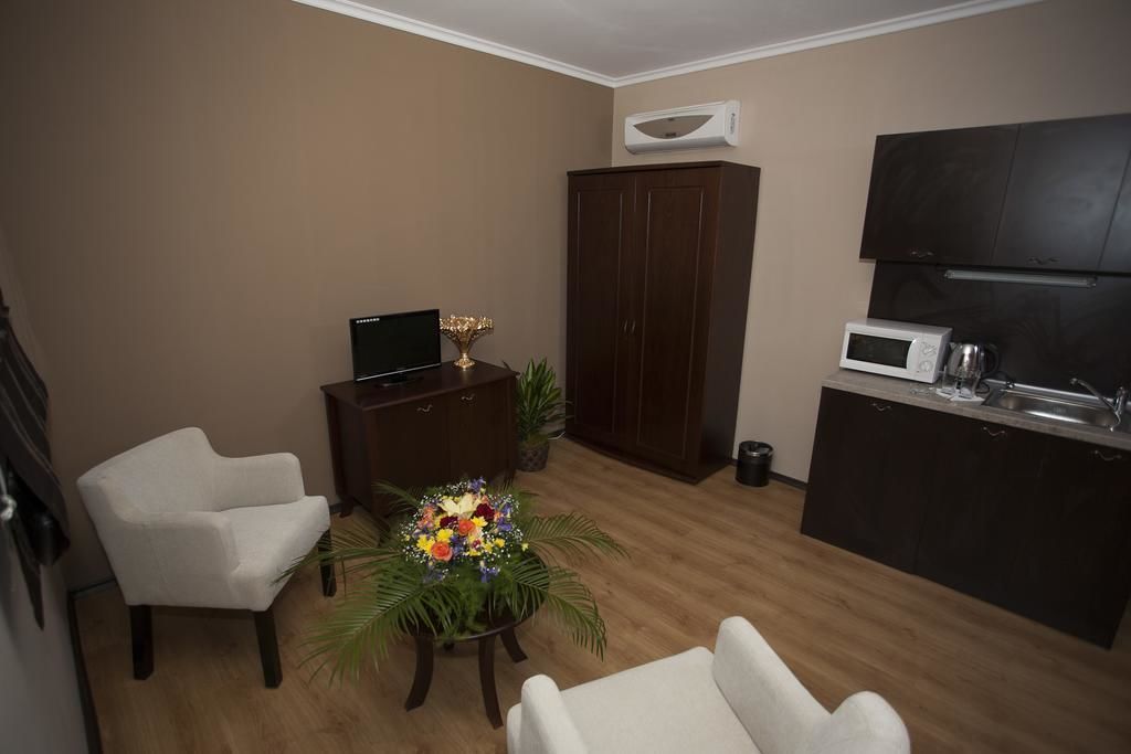 A compact living space with a kitchenette, a dark wooden wardrobe, a white armchair, a small table with a floral arrangement, and a flat-screen TV on a wooden cabinet.
