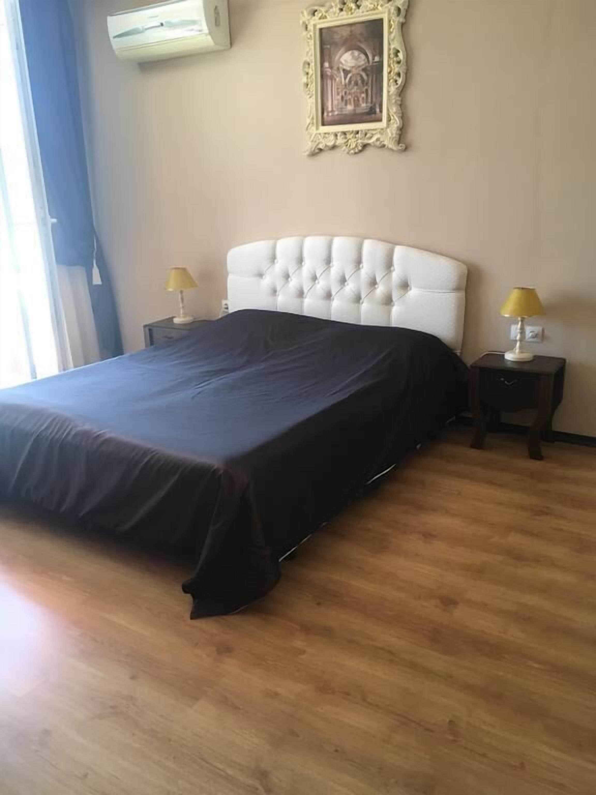 A bedroom with a large bed, two bedside tables with lamps, a framed picture on the wall, and an air conditioner.