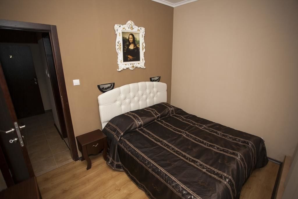 Un dormitorio con un cabecero blanco tapizado, una colcha marrón oscuro, una obra de arte enmarcada en la pared y un banco de madera junto a la cama.