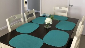 Ein Essbereich mit dunklem Tisch, petrolfarbenen Platzsets, weißen Stühlen, einer kleinen Vase mit Blumen, einem Deckenventilator und einem Schild mit der Aufschrift 'Welcome' an der Wand.