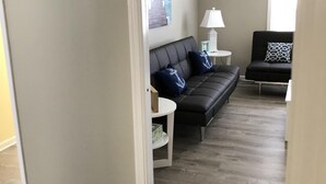 Ein Wohnzimmer mit einem dunkelbraunen Ledersofa, einem weißen Beistelltisch und einem schwarzen Sessel. Eine Wandlampe und ein kleiner Tisch mit einer Lampe sind zu sehen. Auf der Wand steht das Wort 'Beach'.