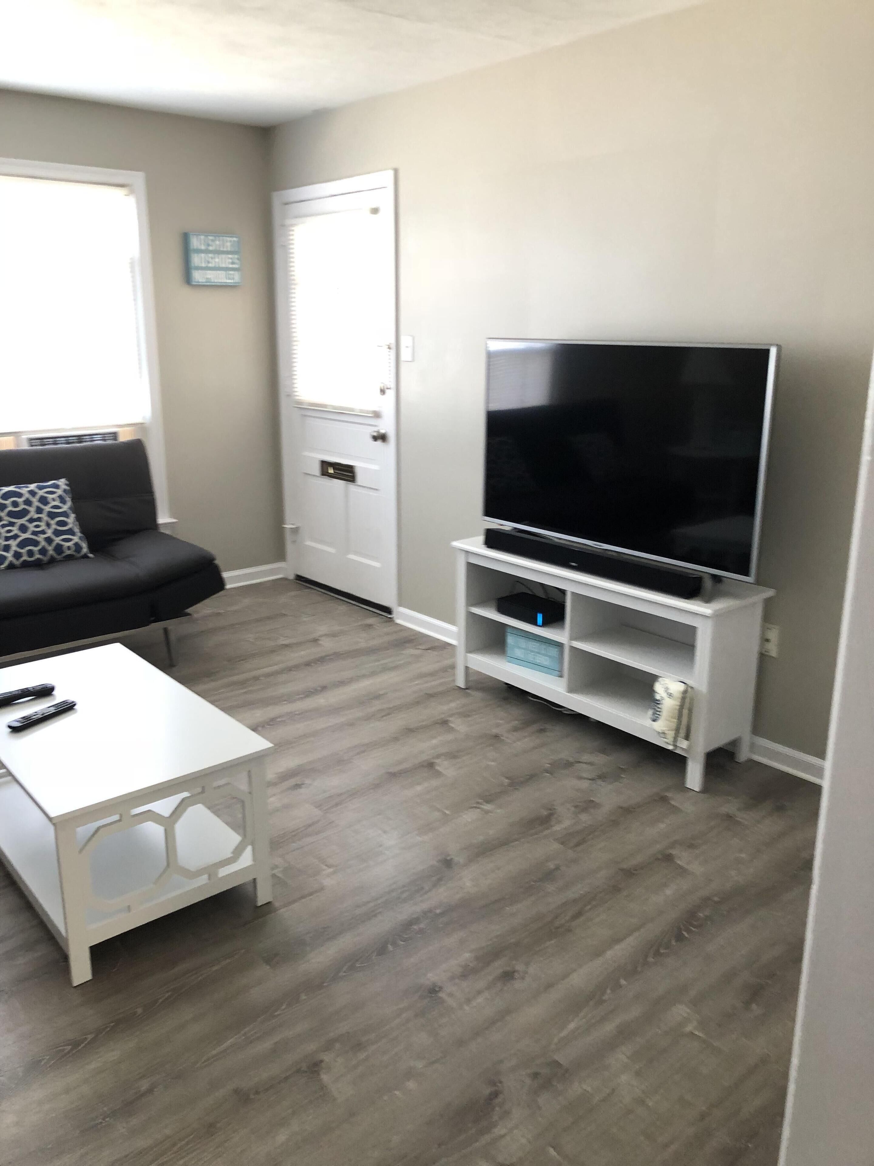 Un salon avec un canapé gris, une table basse blanche, un téléviseur à écran plat posé sur un meuble blanc, et une porte au-dessus de laquelle est accrochée une enseigne.