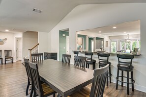 Un comedor amplio con una gran mesa de madera y varias sillas, una zona de cocina con barra, y vistas al exterior a través de amplios ventanales.