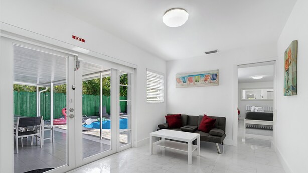 Habitación de hotel moderna con puerta de vidrio que da a una zona de piscina, una mesa de centro blanca y un sofá gris con cojines rojos.