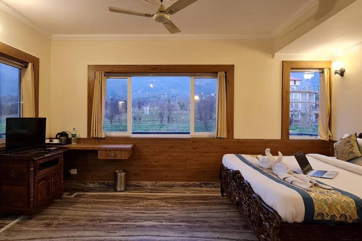 Un dormitorio con una cama grande, un escritorio de madera, un televisor y vistas a un paisaje verde a través de amplios ventanales.
