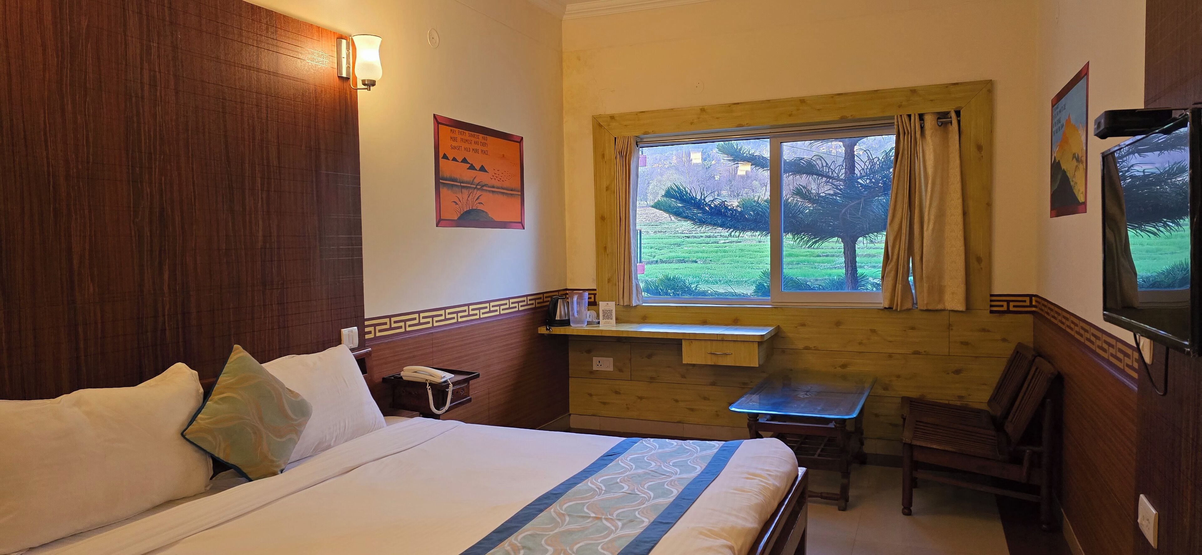 A hotel room with a bed, a desk, a chair, a window with a view of greenery, and a wall-mounted TV.