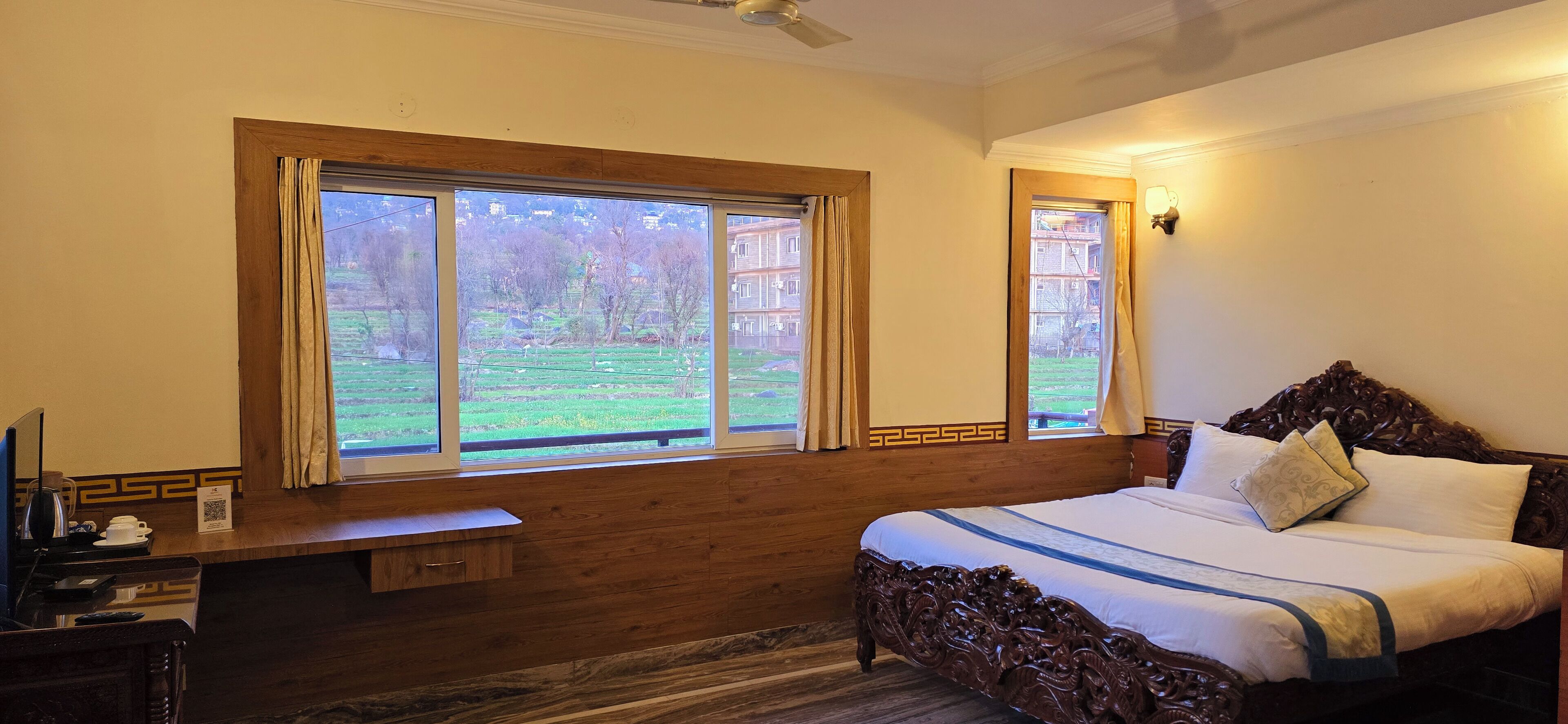 A bedroom with a large window, a wooden bench, a bed with ornate headboard, and a bedside table with a lamp.