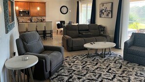 Ein modernes Wohnzimmer mit einer grauen Couch, einem kleinen runden Tisch und einem gemusterten Teppich. Es gibt eine Küchenzeile mit Holzschränken und einer Mikrowelle. Ein Deckenventilator ist sichtbar, und eine Uhr ist an der Wand angebracht.