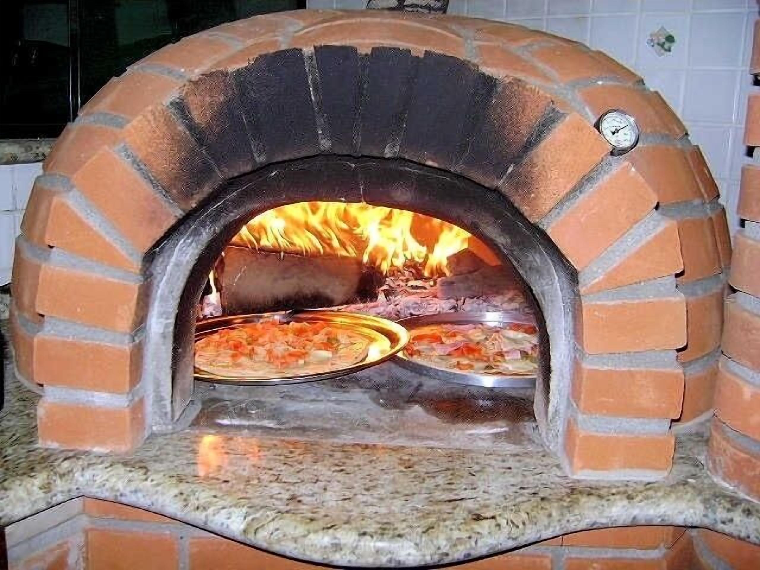 En pizzaugn av tegelsten med två pizzaplattor inuti, en brasa som brinner och en digital termometer på väggen.