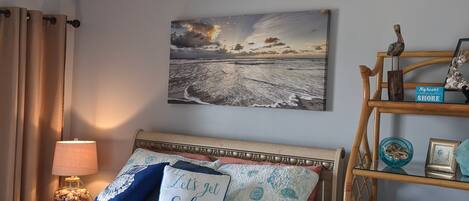 A bedroom with a bed, a wall-mounted painting, a bedside lamp, a wooden shelf with towels, and a decorative bird.