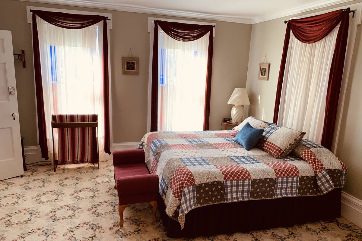 Quarto com uma cama grande, um banco e duas janelas com cortinas.