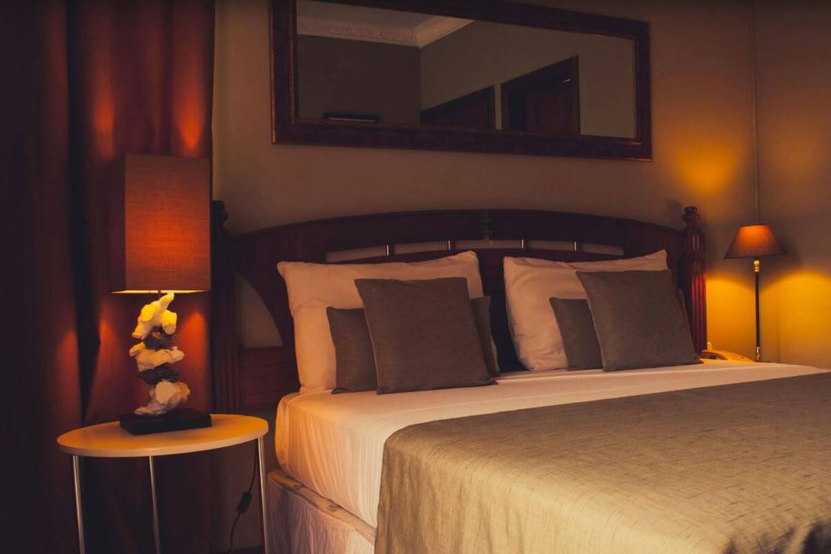Quarto de hotel com cama, duas luminárias de cabeceira, mesa de cabeceira, espelho e luminária fixada na parede.