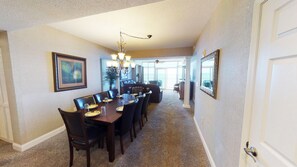 Uma sala de jantar com uma mesa comprida, cadeiras e um lustre. Há uma janela grande com vista para o exterior.