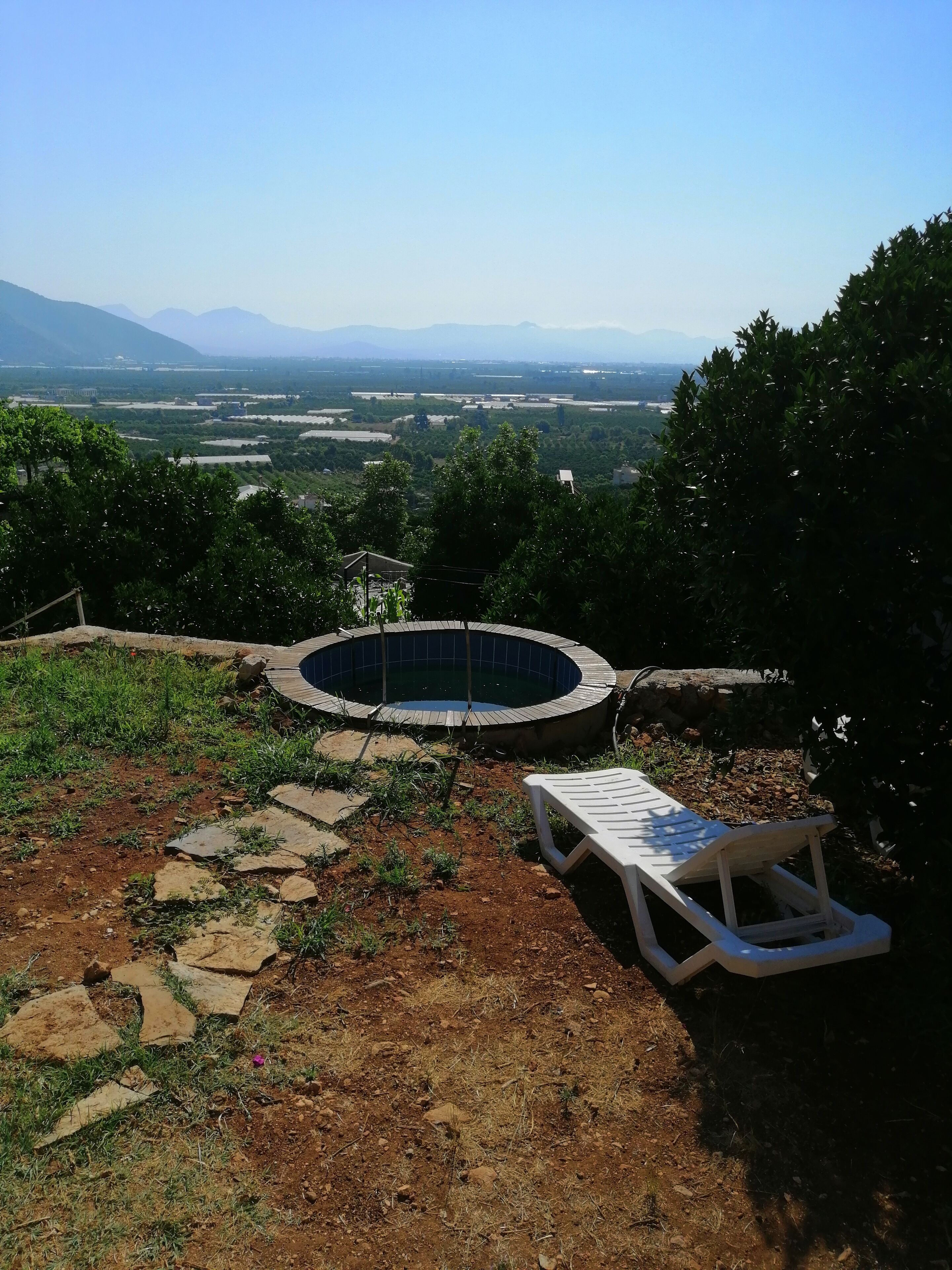 Un jacuzzi rodeado por un sendero de piedra, una silla de lounge de madera, y vistas a montañas y campos lejanos.