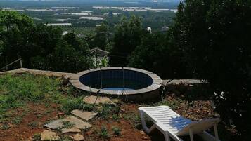 Un jacuzzi rodeado por un sendero de piedra, una silla de lounge de madera, y vistas a montañas y campos lejanos.