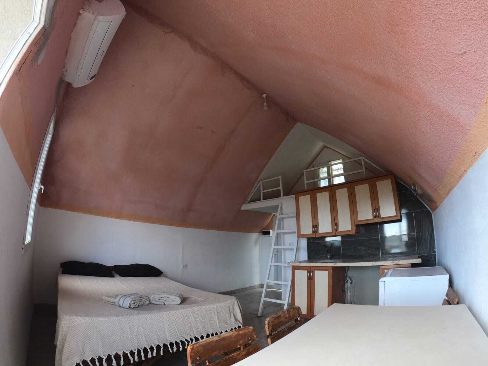 Un dormitorio compacto y triangular en el ático, con cama, una mesita y un rincón de cocina.