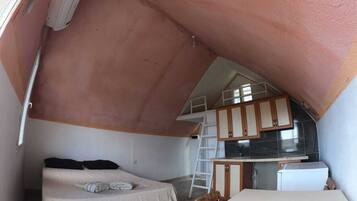 Un dormitorio compacto y triangular en el ático, con cama, una mesita y un rincón de cocina.