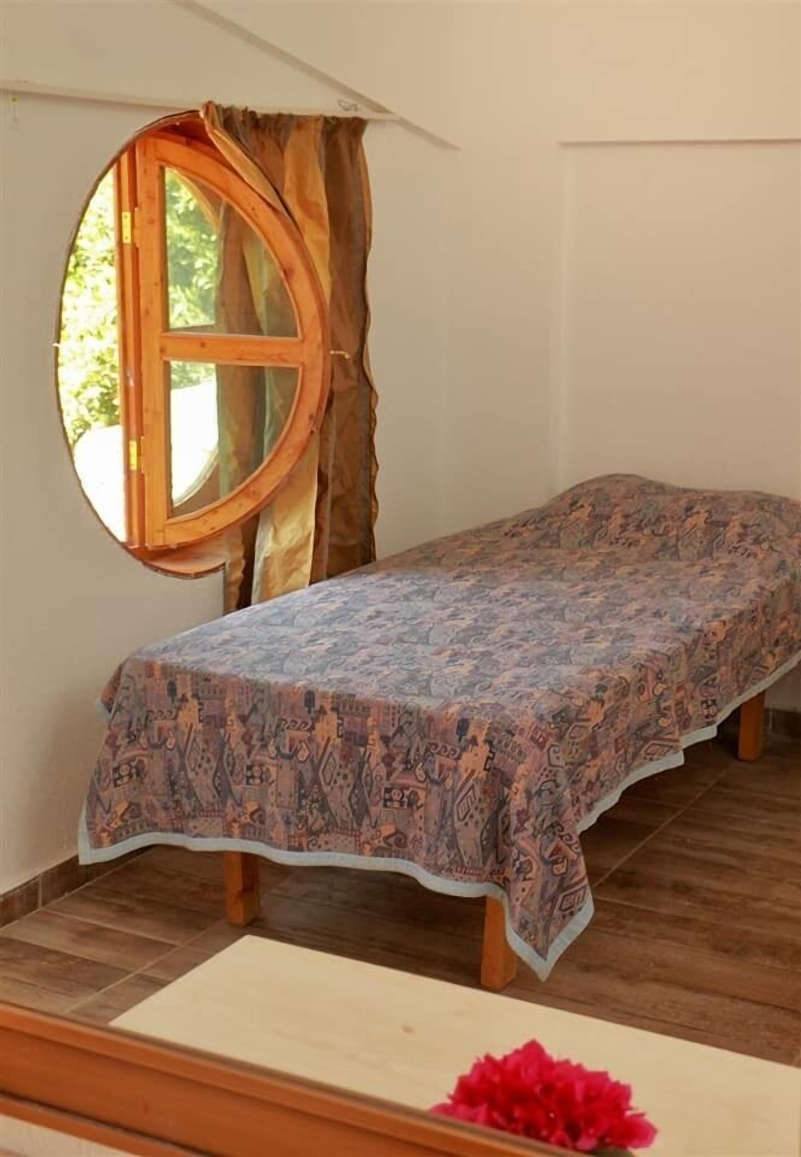 A single bed with a patterned bedspread in a room with a wooden floor and a window with curtains.