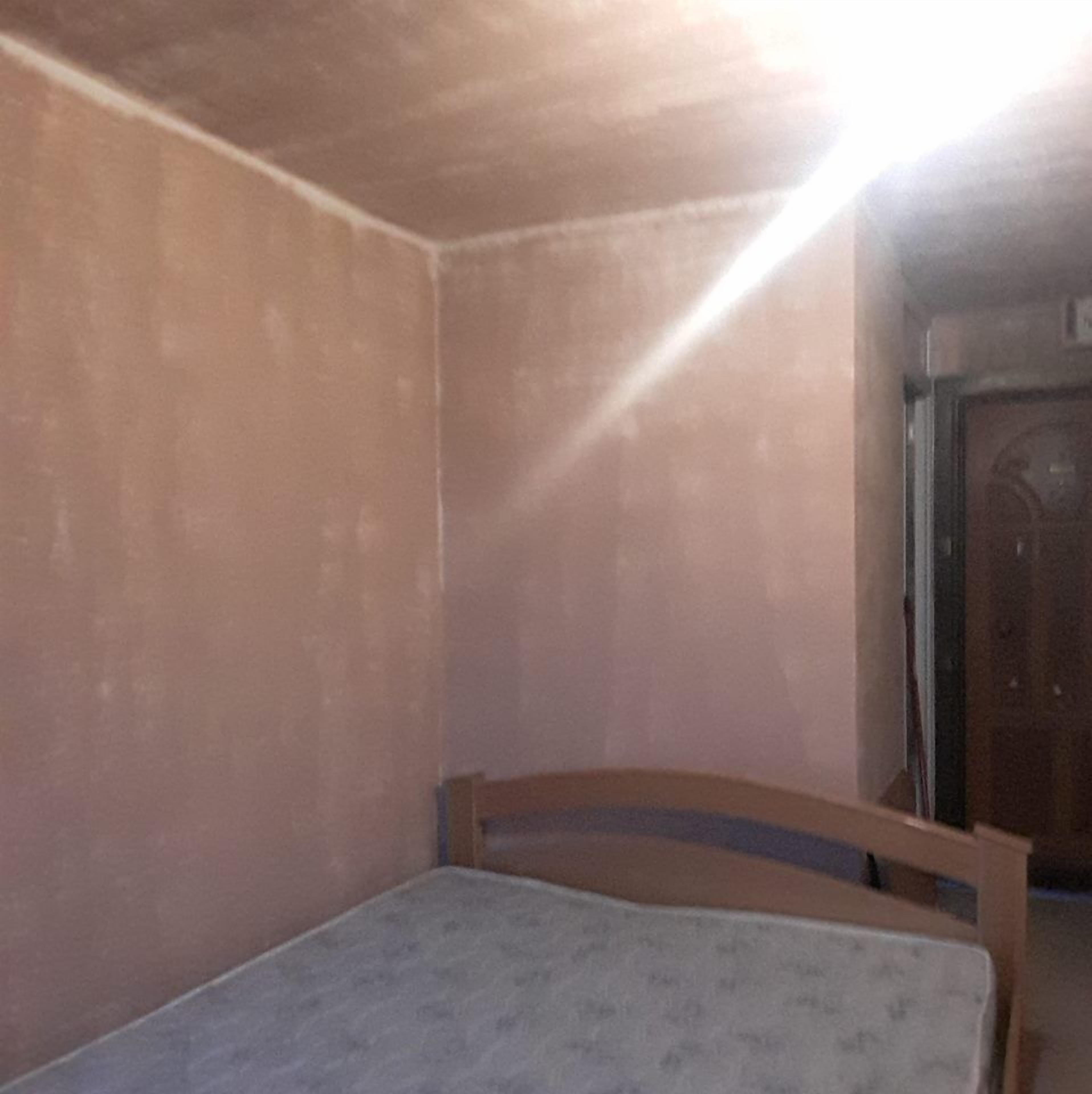 Una habitación pequeña y poco iluminada con una cama individual, paredes lisas y una puerta al fondo.