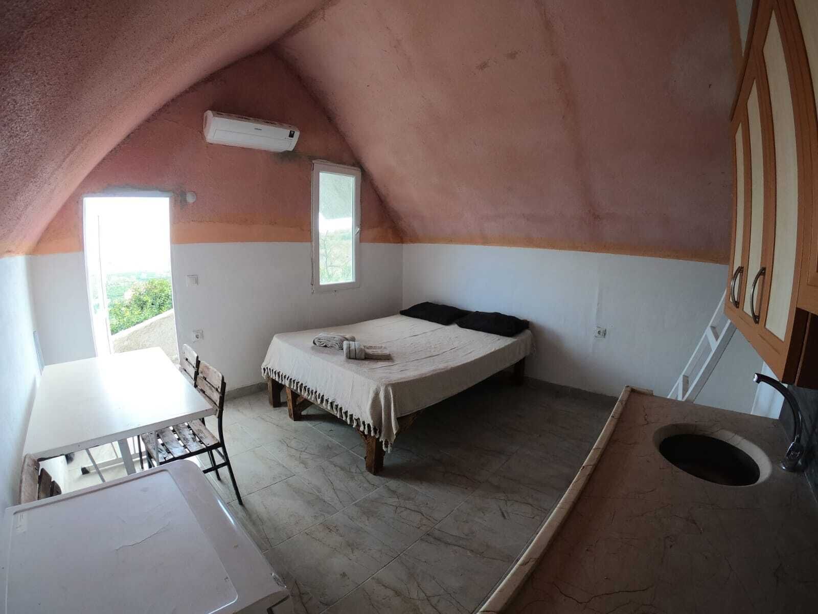 Acomodação pequena, com um único quarto, teto inclinado, cama, mesa pequena e janela.