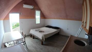 Acomodação pequena, com um único quarto, teto inclinado, cama, mesa pequena e janela.