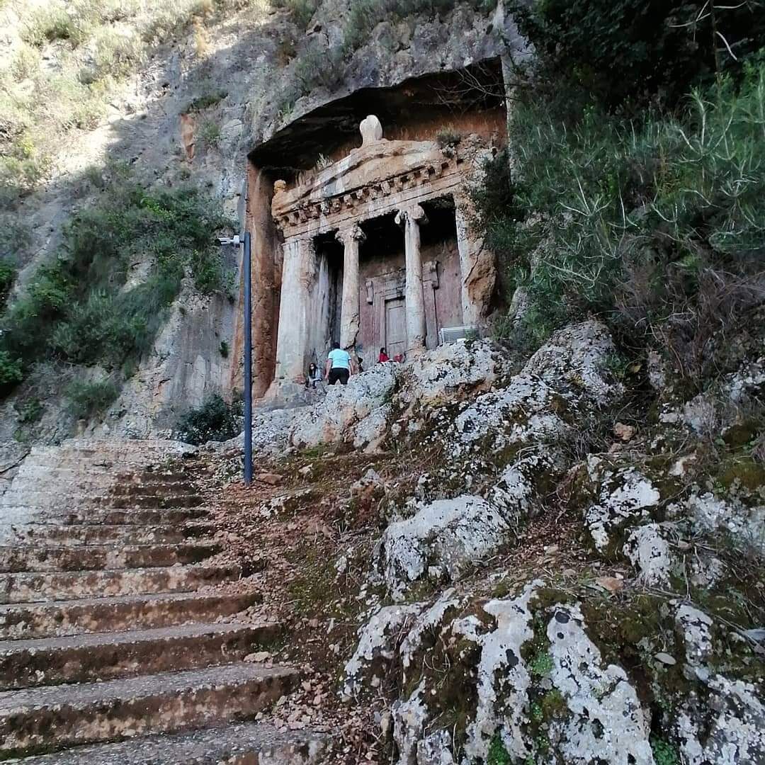 Un escalier en pierre mène à une structure ancienne ornée de colonnes, qui est elle-même entourée d’un terrain rocheux et d’une végétation clairsemée.