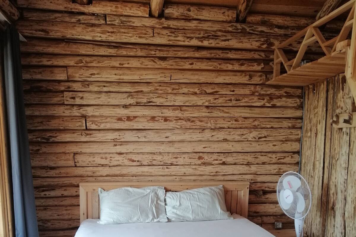 A wooden cabin room with a bed, pillows, and a standing fan.