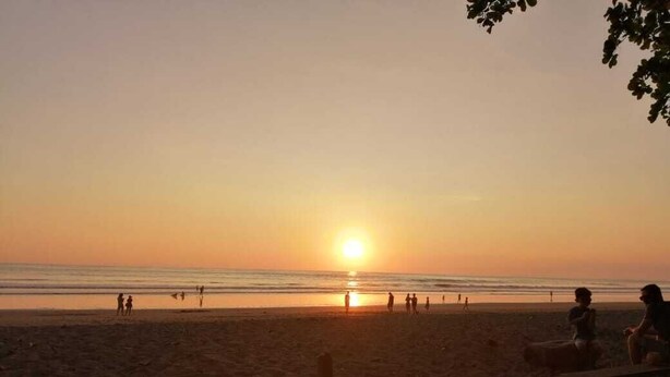 Eine Strandansicht mit untergehender Sonne über dem Meer, Menschen, die am Ufer entlanggehen, und eine hölzerne Bank im Vordergrund.