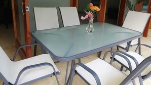 Ein Esstisch mit Glasplatte, umgeben von vier weißen Stühlen mit Metallgestellen. Auf dem Tisch steht eine Vase mit Blumen.