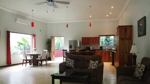 Ein geräumiges Wohnzimmer mit Deckenventilator, ein Essbereich mit Tisch und Stühlen sowie eine Küchenzeile mit Holzschränken und einem Kühlschrank.