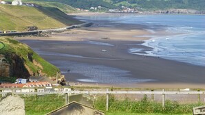 Uma área costeira com uma praia de areia, uma colina gramada e alguns edifícios ao fundo.