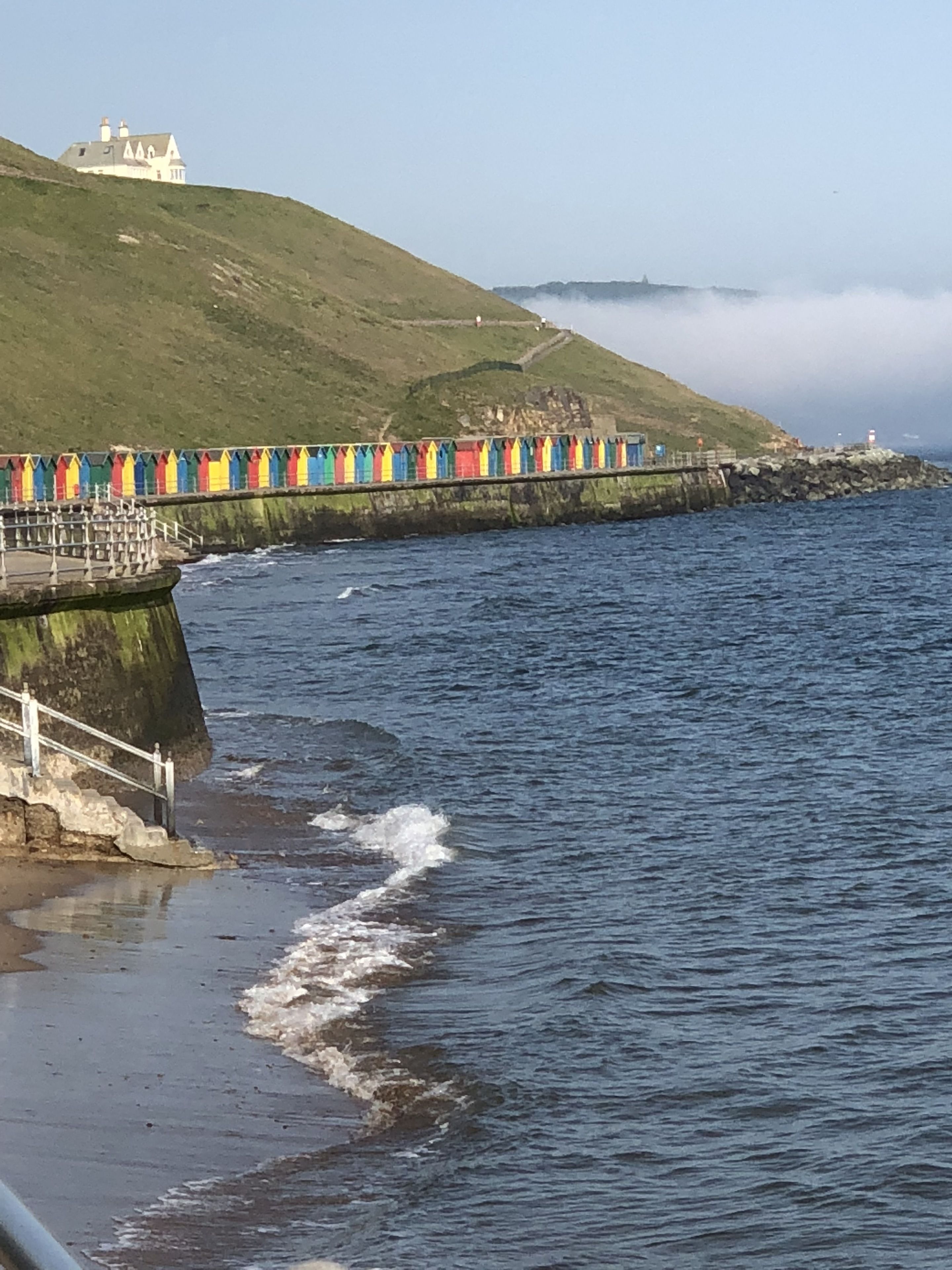 A coastal scene with a building on a hill, a colorful fence, and a rocky shoreline.