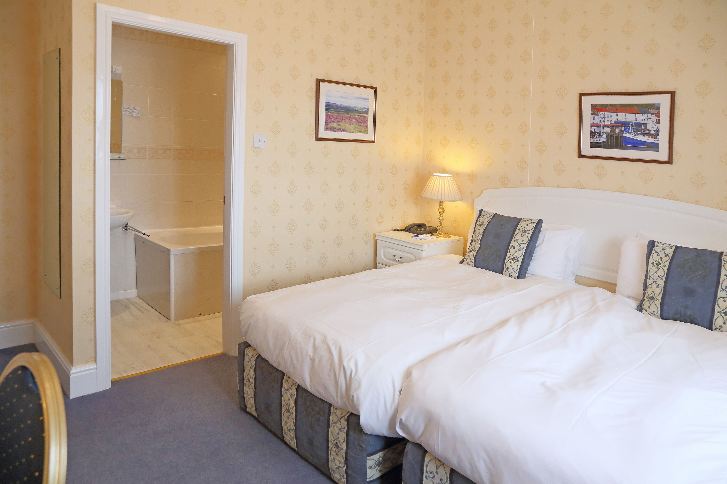 A hotel room with a bed, bedside table, lamp, and a bathroom visible through an open door.