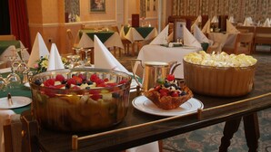 Disposição de buffet com uma grande tigela de frutas, um prato de sobremesas e um jarro de café em uma sala de jantar de hotel.