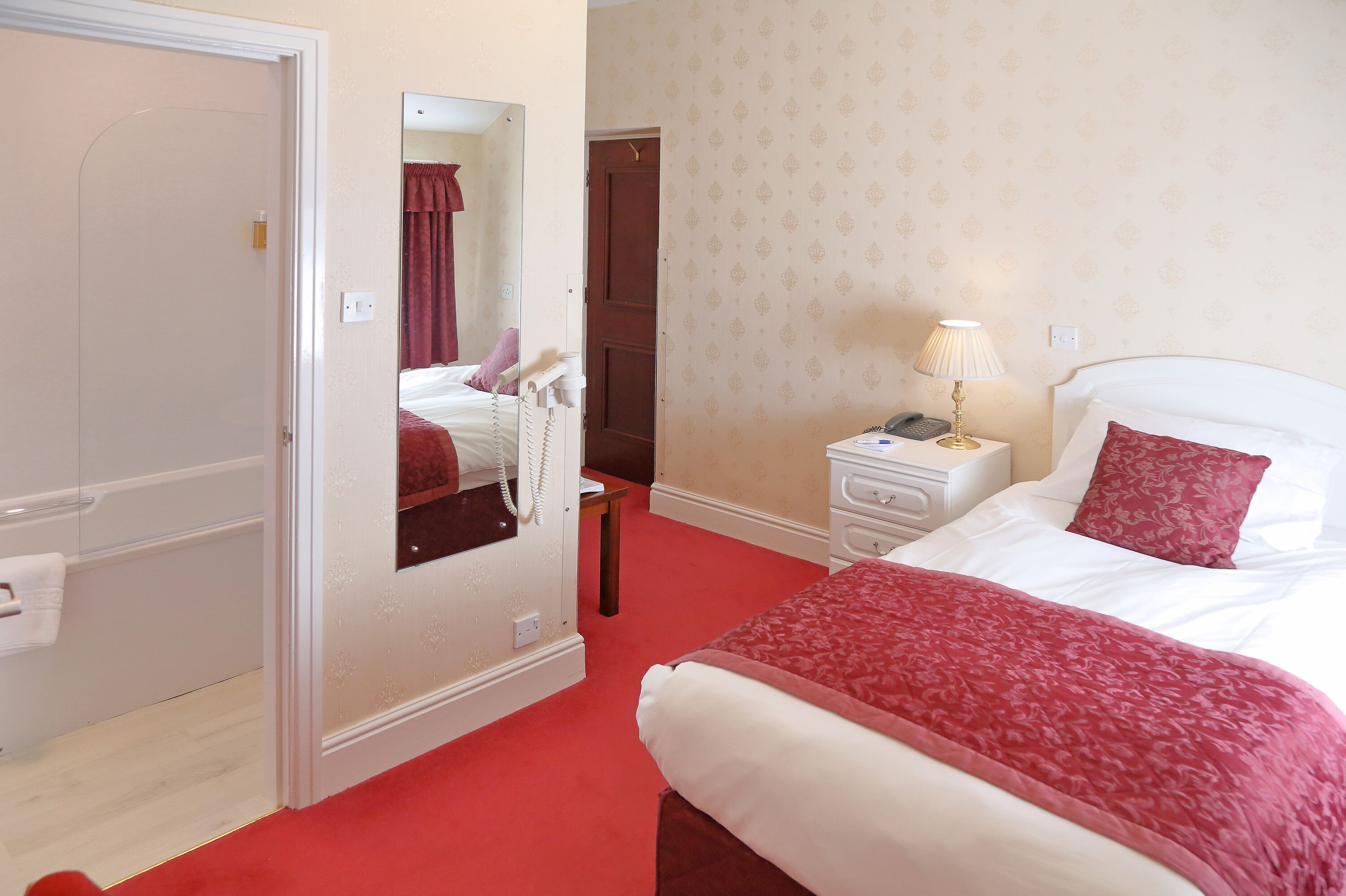 Una habitación de hotel con una cama, una mesita de noche, un espejo, una puerta y una alfombra roja.