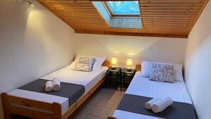 Une petite chambre accueillante avec deux lits en bois, chacun muni d’un oreiller et d’une serviette enroulée. On y trouve deux tables de chevet avec des lampes, et une lucarne au-dessus des lits.