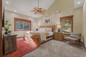 Un dormitorio con cama de madera, mesitas de noche, una silla, ventilador de techo y ventanas con vistas al follaje.