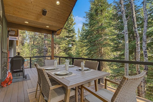 Una terraza de madera al aire libre con una mesa lista para comer, rodeada de sillas y vista a un bosque.