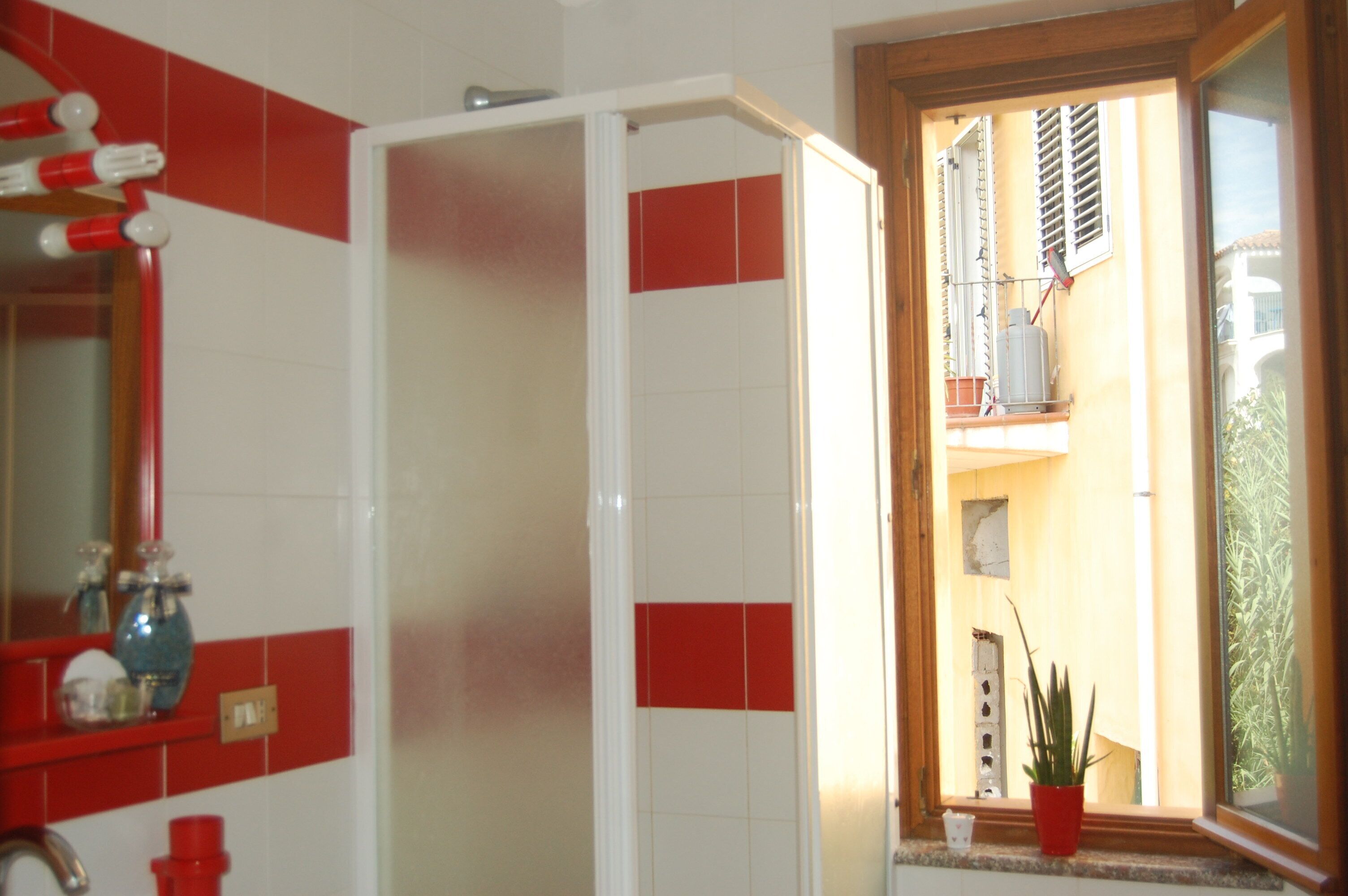 Een badkamer met rode en witte tegels, een douchecabine, een wastafel met rode kraan en een raam met uitzicht op een balkon.