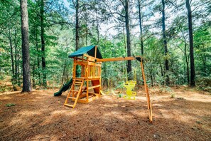 Explore the forest playground with a slide, ladder, and swing set amidst the trees.