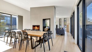 A modern dining area with a large wooden table and black chairs, a fireplace, and a view of the outdoors through large windows.